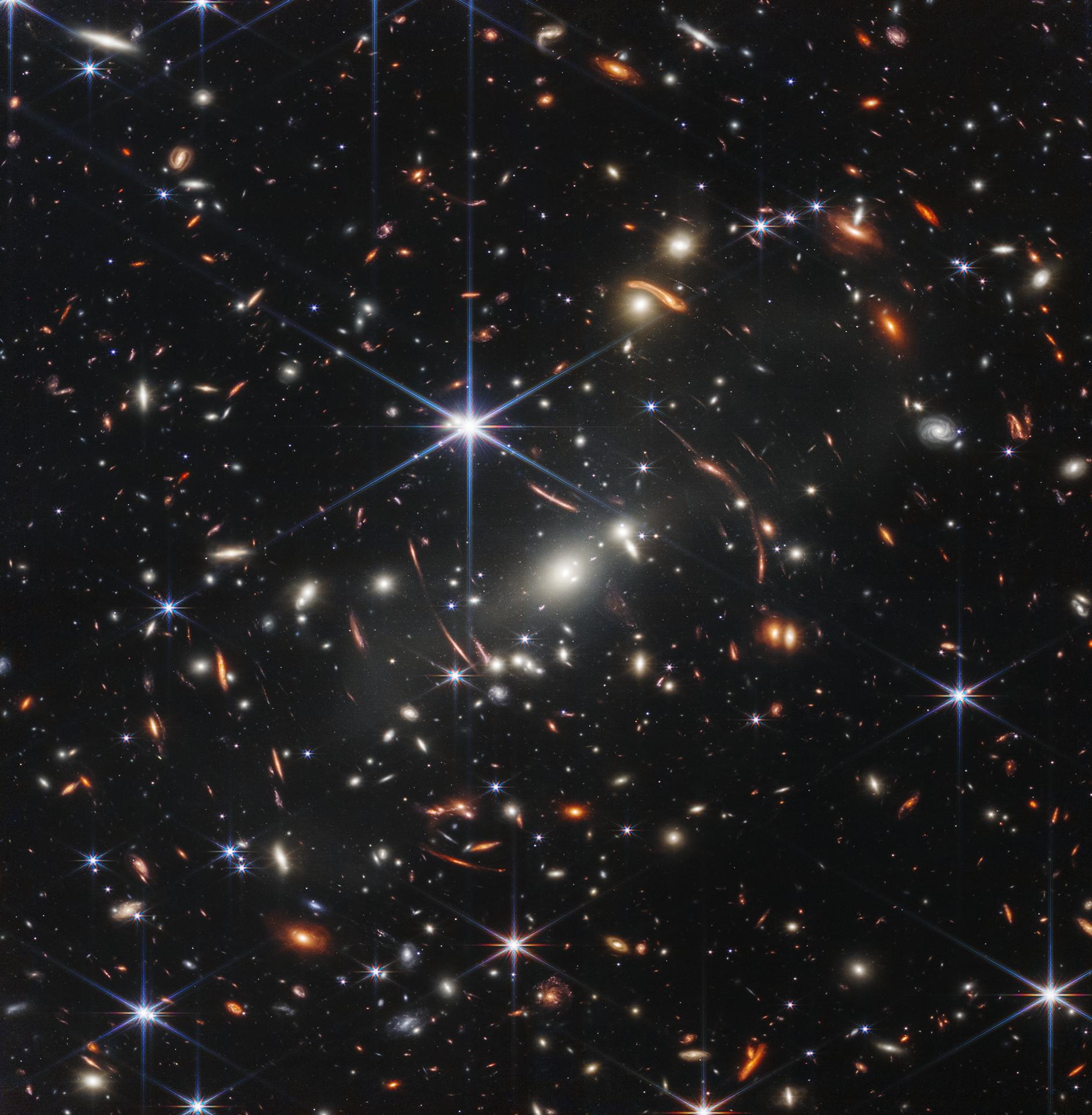 Webb's First Deep Field (SMACS 0723): thousands of galaxies around a foreground galaxy cluster, with lensed galaxies visible as long orange arcs beside the bright central galaxy.
